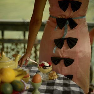 May include: A woman in a pink dress with black bows is standing near a table with a teapot, fruit, and a dessert. The dessert is in a glass bowl and has berries and whipped cream.