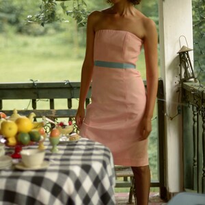 May include: A woman wearing a strapless pink dress with a blue sash stands on a porch with a table set for a meal. The table has a black and white checkered tablecloth and is covered with dishes, fruit, and a teapot.