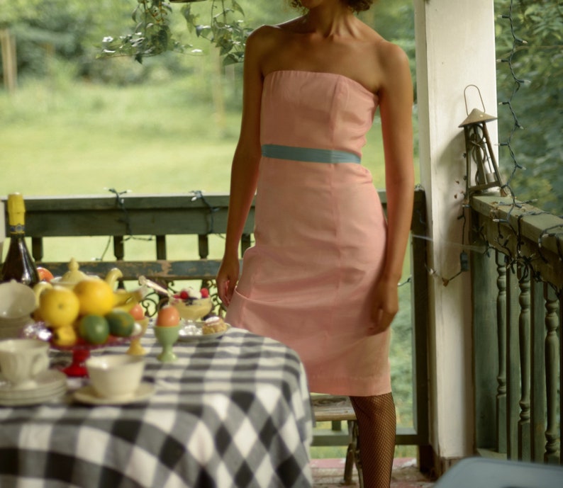 May include: A woman wearing a strapless pink dress with a blue sash stands on a porch with a table set for a meal. The table has a black and white checkered tablecloth and is covered with dishes, fruit, and a teapot.