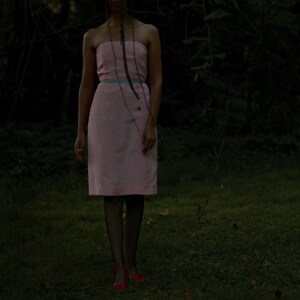 May include: A woman wearing a pink strapless dress with a blue ribbon belt. She is standing in a dark, wooded area.