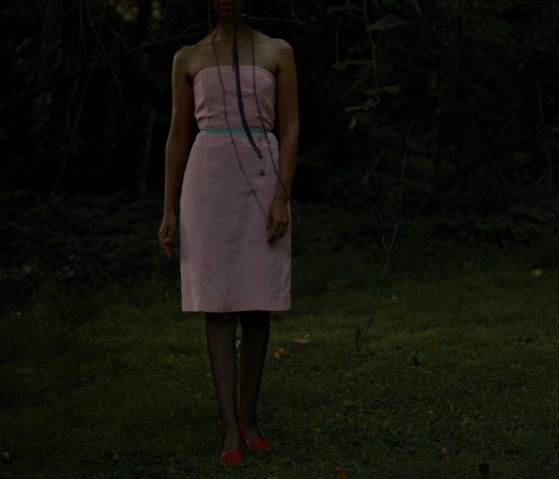 May include: A woman wearing a pink strapless dress with a blue ribbon belt. She is standing in a dark, wooded area.