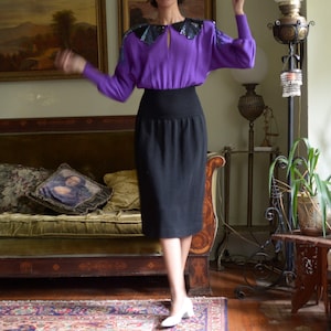 May include: A woman wearing a purple sweater with a sequined collar and a black skirt. She is standing in a room with a sofa, a plant, and a lamp.