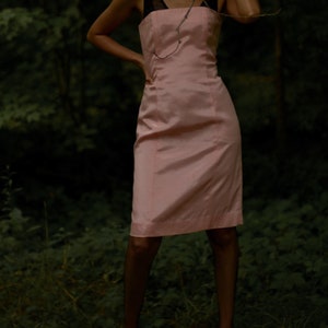 May include: A woman wearing a pink sleeveless dress with a black strap. The dress is fitted and has a straight skirt. The woman is standing in a forest setting.
