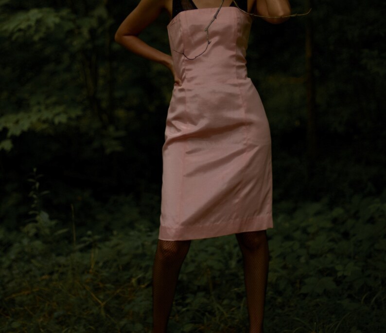 May include: A woman wearing a pink sleeveless dress with a black strap. The dress is fitted and has a straight skirt. The woman is standing in a forest setting.