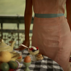 May include: A table setting with a teapot, fruit, eggs, and a dessert with berries and whipped cream. The table has a black and white checkered tablecloth. A person wearing a light pink dress with a blue sash is standing behind the table.