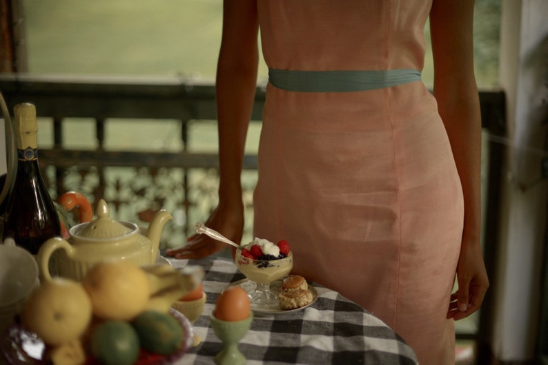 May include: A table setting with a teapot, fruit, eggs, and a dessert with berries and whipped cream. The table has a black and white checkered tablecloth. A person wearing a light pink dress with a blue sash is standing behind the table.