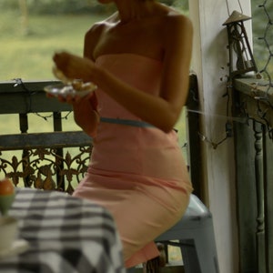 May include: A woman wearing a pink strapless dress sits on a stool on a porch. She is holding a plate of food in her hands.