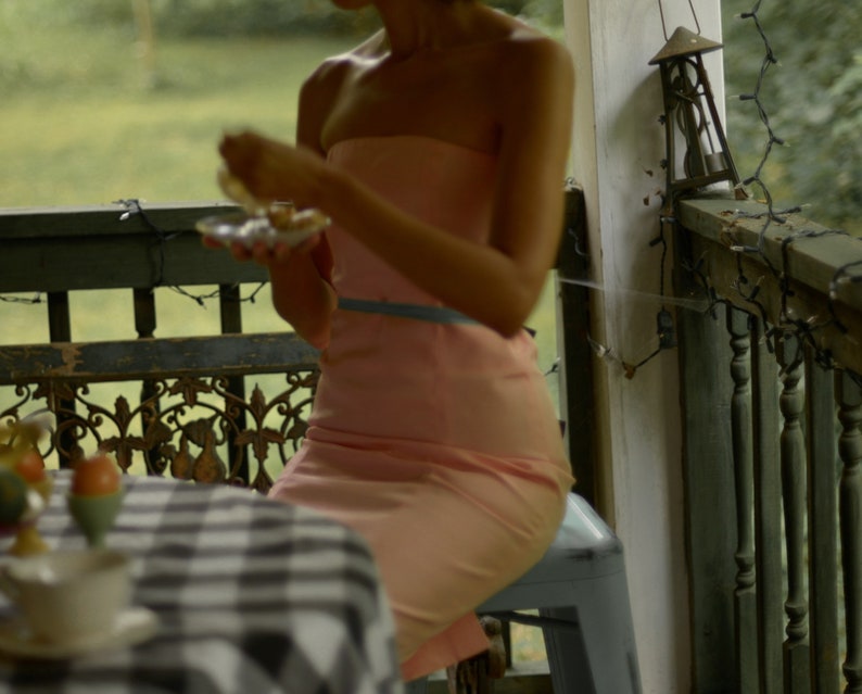 May include: A woman wearing a pink strapless dress sits on a stool on a porch. She is holding a plate of food in her hands.