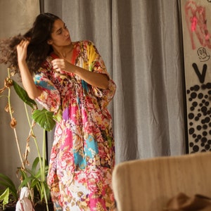 May include: A woman wearing a colorful floral print kaftan dress with a long skirt and short sleeves. She is standing in front of a gray curtain and a wall with graffiti.