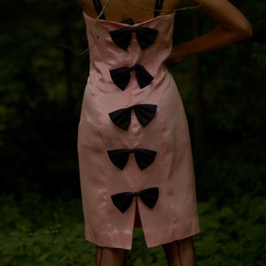 May include: A pink dress with black bow accents on the back. The dress is sleeveless and has a fitted silhouette.