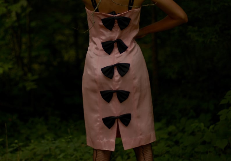 May include: A pink dress with black bow accents on the back. The dress is sleeveless and has a fitted silhouette.