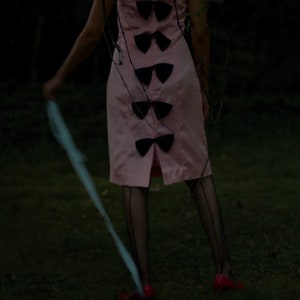 May include: A woman wearing a pink dress with black bows down the back and black fishnet stockings. She is standing in a grassy area with a blue ribbon in her hand.