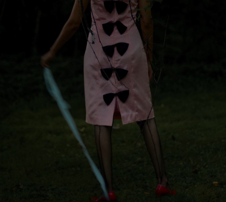 May include: A woman wearing a pink dress with black bows down the back and black fishnet stockings. She is standing in a grassy area with a blue ribbon in her hand.
