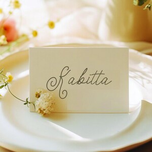 May include: A rectangular off-white place card featuring the name Kabitta written in a dark gray cursive script. A small, dried white flower is attached to the lower left corner of the card, which is positioned on a white plate.