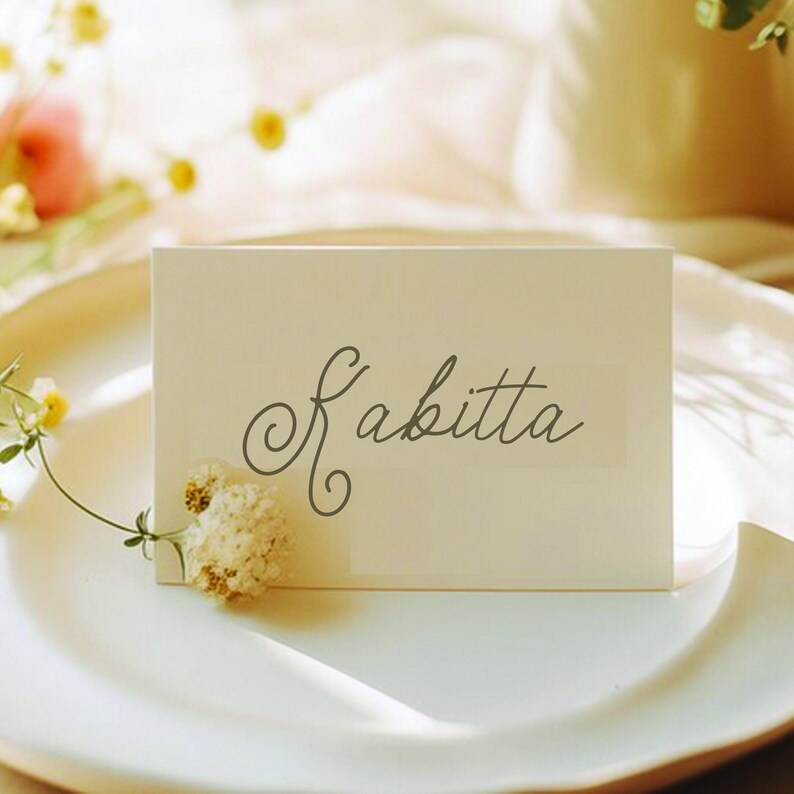 May include: A rectangular off-white place card featuring the name Kabitta written in a dark gray cursive script. A small, dried white flower is attached to the lower left corner of the card, which is positioned on a white plate.