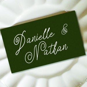 May include: A rectangular dark forest green place card featuring the white cursive text Danielle & Nathan written in a stylized script font. The card is positioned horizontally with a clean, smooth surface and sharp, defined edges.