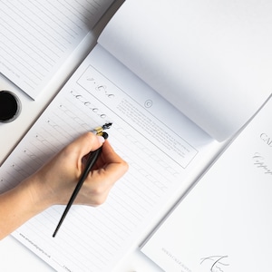 May include: A person's hand writing calligraphy with a black and gold calligraphy pen on a white lined paper pad. The paper pad has the text "www.amaliacaligraphy.co.uk" printed on the bottom. The text "Calligraphy" is printed on the top of the paper pad.