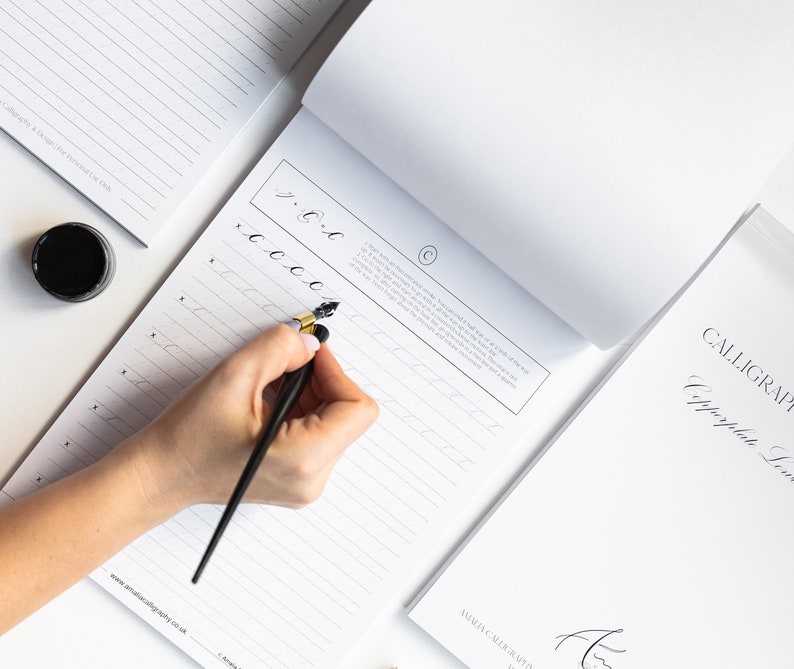 May include: A person's hand writing calligraphy with a black and gold calligraphy pen on a white lined paper pad. The paper pad has the text "www.amaliacaligraphy.co.uk" printed on the bottom. The text "Calligraphy" is printed on the top of the paper pad.
