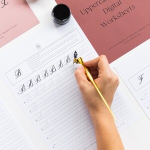 May include: Copperplate calligraphy uppercase letter B practice worksheet with a hand holding a calligraphy pen and ink. The worksheet has a light gray background with a grid pattern and the letter B is written in black ink.