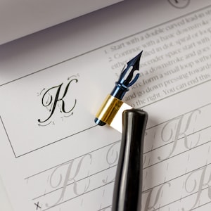 May include: A calligraphy workbook with a blue and gold calligraphy pen and a black calligraphy pen. The workbook has instructions for writing the letter "K" in cursive.