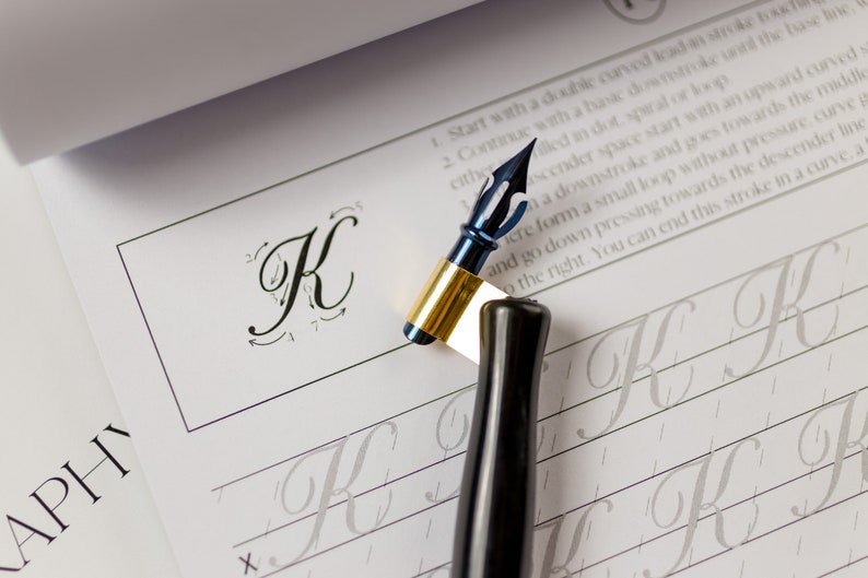 May include: A calligraphy workbook with a blue and gold calligraphy pen and a black calligraphy pen. The workbook has instructions for writing the letter "K" in cursive.