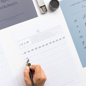 May include: A calligraphy worksheet with instructions on how to write the lowercase letter 'a' in copperplate calligraphy. The worksheet is printed on white paper with gray lines. A person's hand is holding a black calligraphy pen and writing on the worksheet.