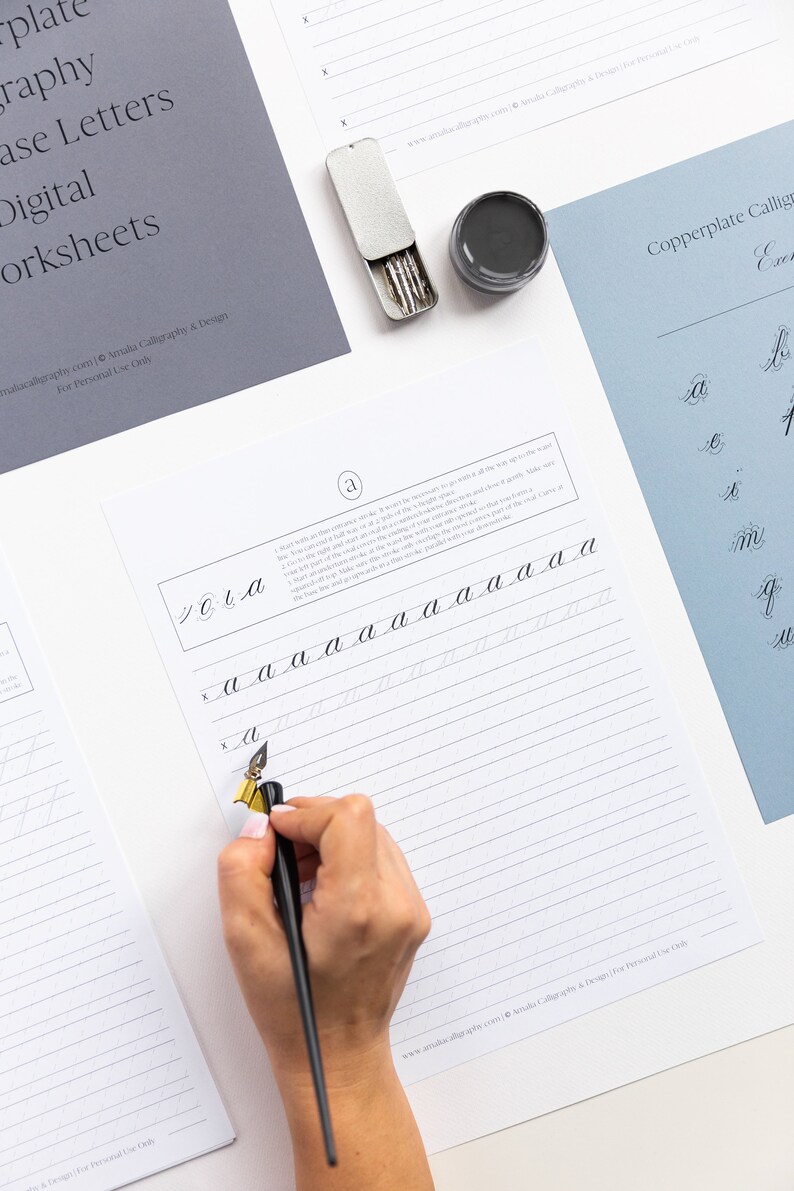 May include: A calligraphy worksheet with instructions on how to write the lowercase letter 'a' in copperplate calligraphy. The worksheet is printed on white paper with gray lines. A person's hand is holding a black calligraphy pen and writing on the worksheet.