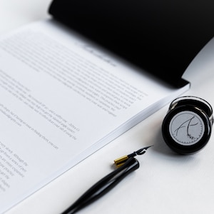 May include: A black inkwell with a white label featuring the letter "A" in a cursive font. A black and gold calligraphy pen is also pictured.