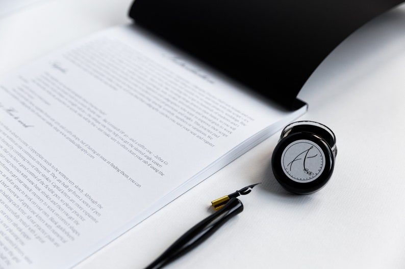 May include: A black inkwell with a white label featuring the letter "A" in a cursive font. A black and gold calligraphy pen is also pictured.