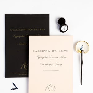 May include: Two calligraphy practice pads, one black and one peach, with the text "Calligraphy Practice Pad" and "Copperplate Lowercase Letters" in gold lettering. The pads are on a white surface with two ink pots and a calligraphy pen.