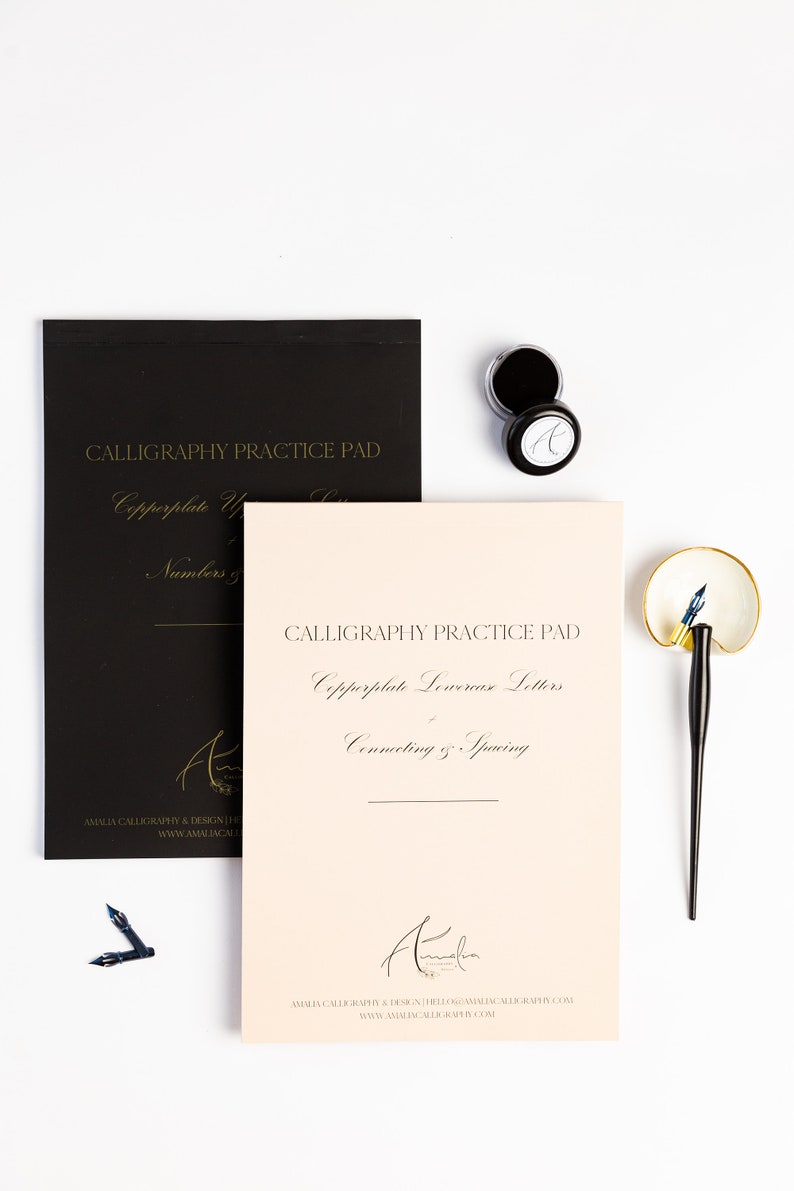 May include: Two calligraphy practice pads, one black and one peach, with the text "Calligraphy Practice Pad" and "Copperplate Lowercase Letters" in gold lettering. The pads are on a white surface with two ink pots and a calligraphy pen.