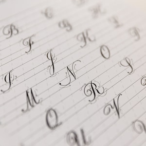 May include: A close-up of a page from a calligraphy practice book. The letters A through W are written in cursive script on lined paper.