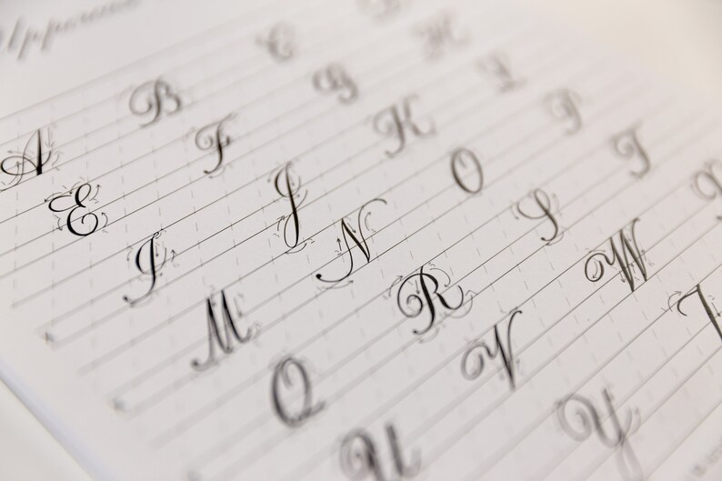 May include: A close-up of a page from a calligraphy practice book. The letters A through W are written in cursive script on lined paper.