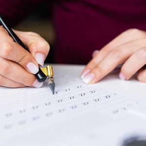 May include: A person's hand holding a black and gold calligraphy pen and writing on a lined sheet of paper. The pen has a nib and a black handle with a gold band. The person is practicing calligraphy by writing the letter 'd' repeatedly.