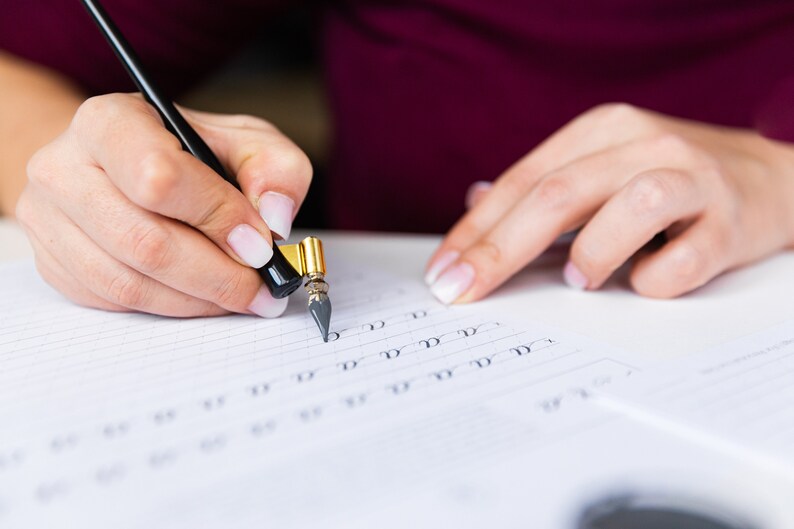 May include: A person's hand holding a black and gold calligraphy pen and writing on a lined sheet of paper. The pen has a nib and a black handle with a gold band. The person is practicing calligraphy by writing the letter 'd' repeatedly.