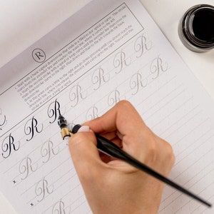 May include: A person practicing calligraphy with a black calligraphy pen and ink on a lined paper pad. The paper has instructions for writing the letter "R" in cursive. The instructions include starting with a basic capital downstroke that ends in a filled-in spiral or curve.