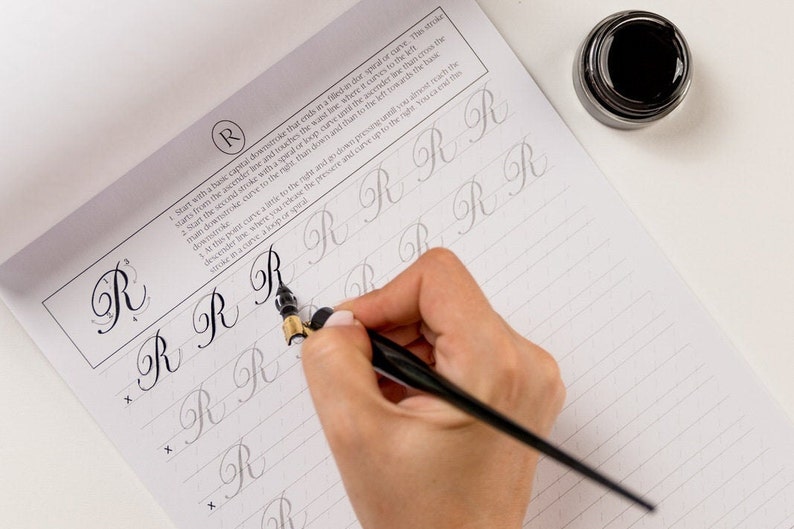 May include: A person practicing calligraphy with a black calligraphy pen and ink on a lined paper pad. The paper has instructions for writing the letter "R" in cursive. The instructions include starting with a basic capital downstroke that ends in a filled-in spiral or curve.