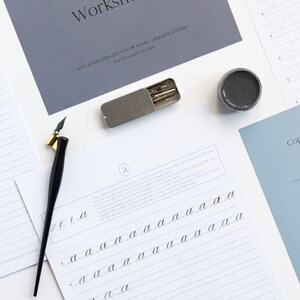 May include: A calligraphy worksheet for practicing lowercase letter 'a' with instructions and multiple lines for writing. The worksheet is on white paper with gray lines. A black calligraphy pen, a gray ink pot, and a silver case with calligraphy nibs are also in the image.