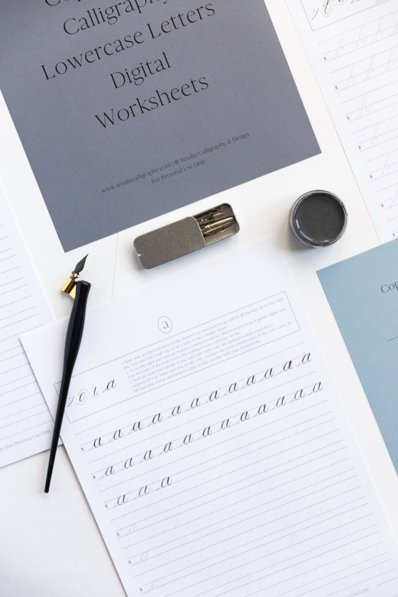 May include: A calligraphy worksheet for practicing lowercase letter 'a' with instructions and multiple lines for writing. The worksheet is on white paper with gray lines. A black calligraphy pen, a gray ink pot, and a silver case with calligraphy nibs are also in the image.