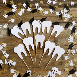 May include: White tooth-shaped cupcake toppers, some wearing black graduation caps, arranged on wooden sticks. Scattered around are gold star and tooth-shaped confetti, and black graduation cap confetti. The theme is dental school graduation.