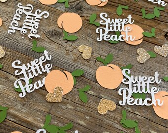 May include: Peach-shaped confetti with the text "Sweet Little Peach" in white lettering. The confetti is on a wooden surface with green leaves and gold glitter hearts.