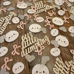 May include: A collection of baby shower decorations on a wooden surface. The decorations include dumpling-shaped cutouts with smiling faces, pink bows, and silver glitter hearts. The phrase "our little dumpling" is also present.