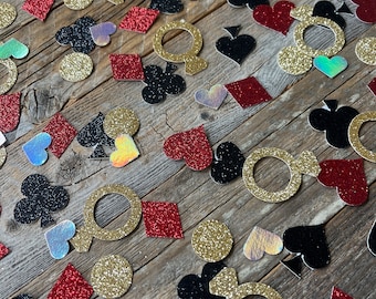 May include: A scattering of red, black, gold, and iridescent glitter confetti featuring various playing card symbols, including hearts, diamonds, spades, and clubs, as well as rings.