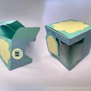 May include: Two teal mug boxes with yellow cloud-shaped accents. One box is open, revealing a clear window for the mug. The boxes have a dotted pattern and a label that says "Mug Box" with a mug graphic. The boxes are designed for gift-giving.