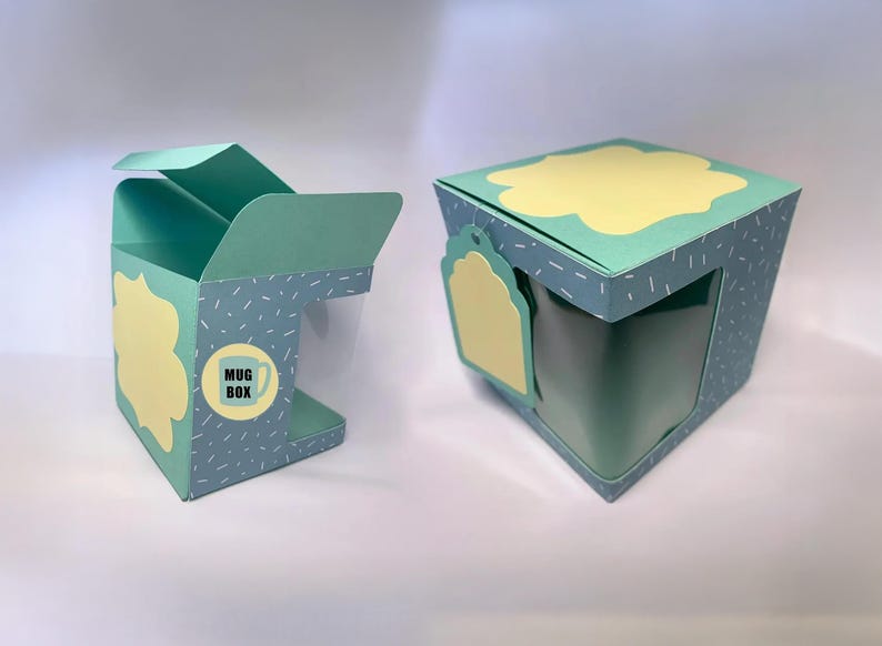 May include: Two teal mug boxes with yellow cloud-shaped accents. One box is open, revealing a clear window for the mug. The boxes have a dotted pattern and a label that says "Mug Box" with a mug graphic. The boxes are designed for gift-giving.