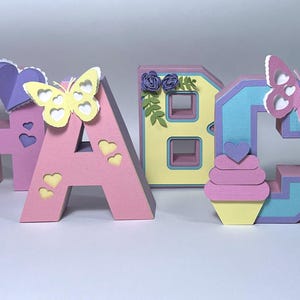 以下が含まれることがあります： ピンク、黄色、青色の紙細工の文字A、B、C。ハートと蝶の装飾が施されています。文字Aには黄色の蝶とハートの切り抜きがあります。文字Bには花の模様があります。文字Cにはピンクの蝶とカップケーキがあります。