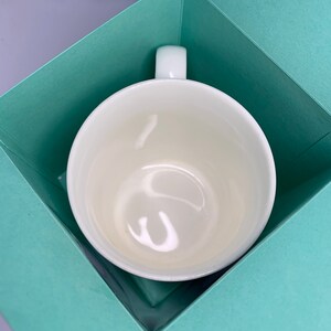 May include: A white ceramic mug with a rounded bowl and a rectangular handle, presented inside a light teal gift box. The mug has a glossy finish and is centered in the box, which is open and angled.