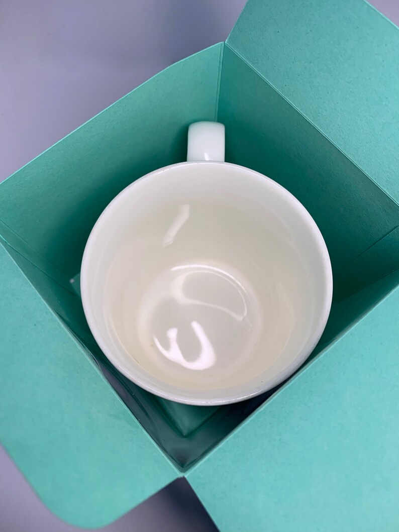May include: A white ceramic mug with a rounded bowl and a rectangular handle, presented inside a light teal gift box. The mug has a glossy finish and is centered in the box, which is open and angled.