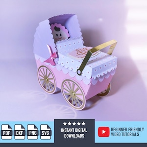 May include: A pink and white paper baby carriage with gold detailing. The carriage features a scalloped hood and wheels, and a small baby figure is visible inside. A gold sign reads "Baby Girl".