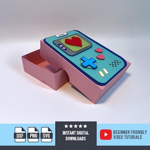 May include: A pink paper box shaped like a retro handheld game console, with a teal top featuring a red heart, a blue cross, and pink hearts. The box is open, revealing the interior. The image includes icons for digital downloads and video tutorials.
